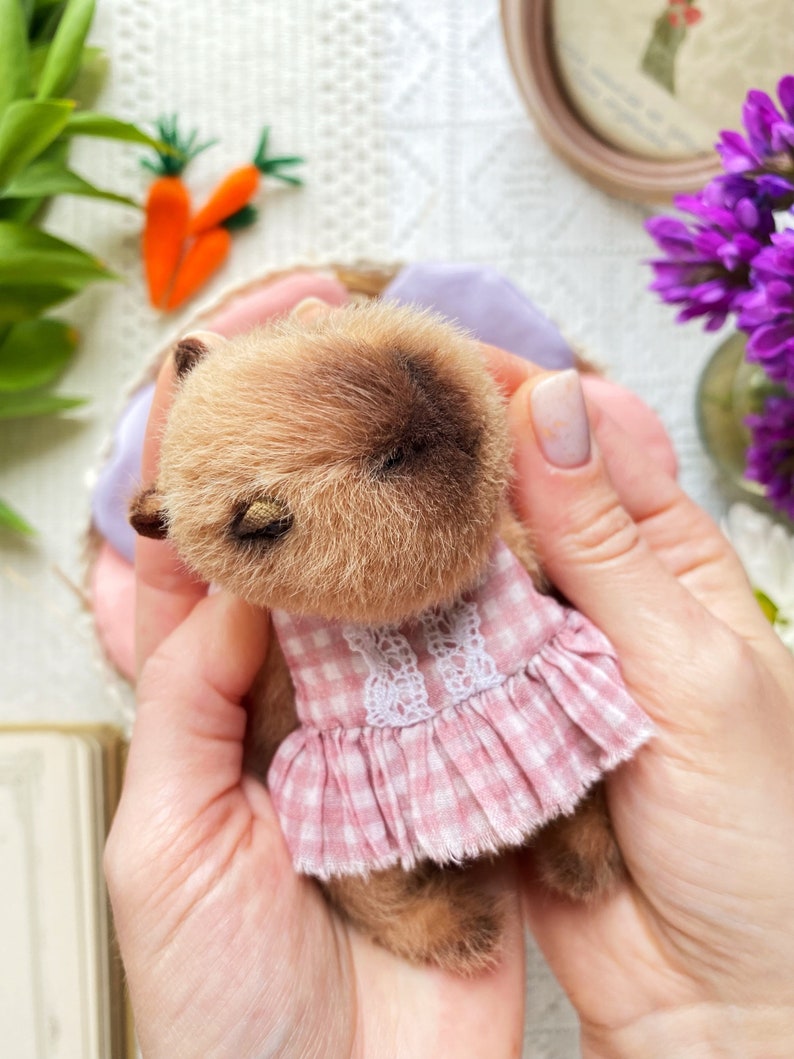 K&ouml;nnte beinhalten: Ein handgefertigter kleiner Teddyb&auml;r mit braunem Fell und einem rosa Vichy-Kleid mit wei&szlig;er Spitzenborte. Der B&auml;r hat geschlossene Augen und wird in H&auml;nden gehalten. Im Hintergrund sind Karotten und lila Blumen zu sehen.