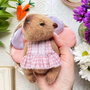 K&ouml;nnte beinhalten: Ein handgefertigter brauner Teddyb&auml;r mit einem rosa Vichy-Kleid mit wei&szlig;er Spitzenborte. Der B&auml;r h&auml;lt ein rosa-lila bl&uuml;tenf&ouml;rmiges Kissen. Zwei orangefarbene Filzkarotten mit gr&uuml;nen Spitzen sind im Hintergrund.