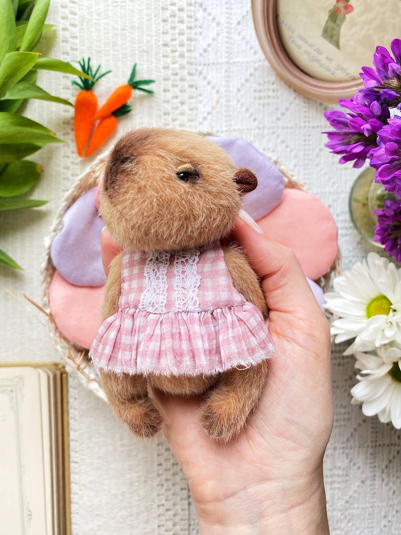 K&ouml;nnte beinhalten: Ein handgefertigter brauner Teddyb&auml;r mit einem rosa Vichy-Kleid mit wei&szlig;er Spitzenborte. Der B&auml;r h&auml;lt ein rosa-lila bl&uuml;tenf&ouml;rmiges Kissen. Zwei orangefarbene Filzkarotten mit gr&uuml;nen Spitzen sind im Hintergrund.