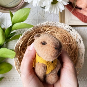 K&ouml;nnte beinhalten: Eine handgefertigte Miniatur-Capybara-Figur mit weichem, hellbraunem Fell. Das kleine Tier tr&auml;gt eine gelbe Weste mit schwarzen Punkten. Es wird in geformten H&auml;nden gehalten, eingebettet in einem geflochtenen Korb.