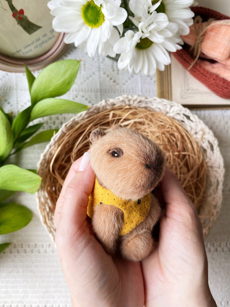 K&ouml;nnte beinhalten: Eine handgefertigte Miniatur-Capybara-Figur mit weichem, hellbraunem Fell. Das kleine Tier tr&auml;gt eine gelbe Weste mit schwarzen Punkten. Es wird in geformten H&auml;nden gehalten, eingebettet in einem geflochtenen Korb.