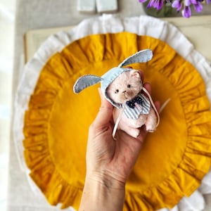 May include: A small, pink teddy bear wearing a light blue bunny hat with white polka dots and a striped bow tie. The bear is held over a yellow, ruffled surface. A bouquet of purple flowers and decorative houses are in the background.