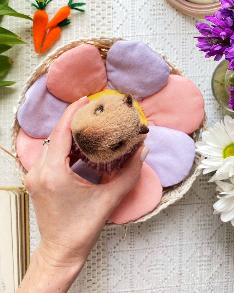 K&ouml;nnte beinhalten: Ein hellbraunes Meerschweinchen-Spielzeug mit gelbem Kopf, eingebettet in einem Korb mit pastellfarbenen Bl&uuml;tenbl&auml;ttern. Der Korb ist von Filzkarotten und Blumen umgeben, was ein skurriles Fr&uuml;hlingsmotiv ergibt.