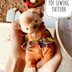 Puede incluir: Un patrón de costura PDF para un adorable juguete de peluche de gato hecho a mano. El gato lleva un mono marrón con estampado floral y una aplicación de oso marrón. El patrón está disponible para descargar.