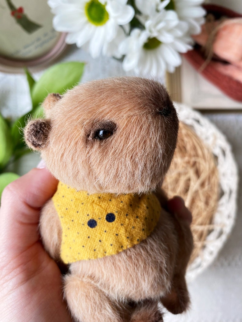 K&ouml;nnte beinhalten: Ein handgefertigtes, miniaturisiertes Capybara-Pl&uuml;schtier mit hellbraunem Fell und einer gelben Weste mit schwarzen Punkten. Das Pl&uuml;schtier hat dunkle Augen und kleine Ohren. Das Spielzeug wird in einer Hand gehalten, mit einem verschwommenen Hintergrund aus wei&szlig;en Blumen und Gr&uuml;n.