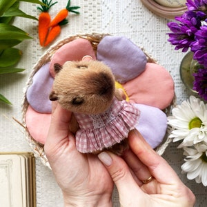 K&ouml;nnte beinhalten: Ein handgefertigter brauner Teddyb&auml;r mit einem rosa Vichy-Kleid mit wei&szlig;er Spitzenborte. Der B&auml;r liegt in einem bl&uuml;tenf&ouml;rmigen Korb mit rosa und lila Bl&uuml;tenbl&auml;ttern. Filzkarotten und Blumen sind im Hintergrund.