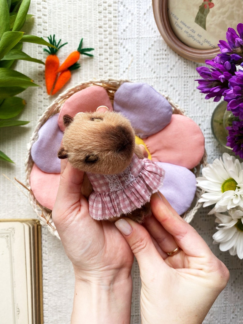 K&ouml;nnte beinhalten: Ein handgefertigter brauner Teddyb&auml;r mit einem rosa Vichy-Kleid mit wei&szlig;er Spitzenborte. Der B&auml;r liegt in einem bl&uuml;tenf&ouml;rmigen Korb mit rosa und lila Bl&uuml;tenbl&auml;ttern. Filzkarotten und Blumen sind im Hintergrund.