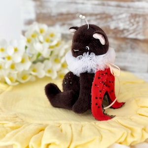 Può includere: Una bambola di gatto di peluche marrone fatta a mano con un colletto bianco a balze e un accessorio rosso a tema coccinella. La bambola ha piccole orecchie, un viso delicato ed è adornata con minuscoli abbellimenti.