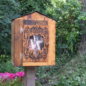 Puede incluir: Una pequeña biblioteca gratuita de madera con una puerta decorativa tallada. La puerta tiene una ventana de cristal y un borde oscuro y adornado. La biblioteca está montada sobre un poste de madera y rodeada de vegetación y flores rosas.