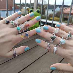 Op de afbeelding: Een verzameling ringen met verschillende halfedelstenen die zijn gewikkeld in zilverdraad. De stenen omvatten amethist, aquamarijn, rozenkwarts en meer.