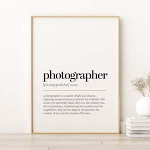 Pode incluir: Uma definição imprimível em preto e branco da palavra "fotógrafo" com a ortografia fonética e uma descrição do que um fotógrafo faz. A definição está emoldurada em uma moldura dourada e está sobre uma superfície de madeira.