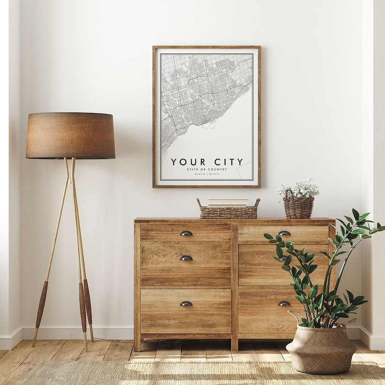 May include: A black and white city map print in a wooden frame with the text "YOUR CITY, STATE OR COUNTRY" hanging above a wooden dresser with three drawers on each side. The dresser is in a room with a white wall, a brown floor, a brown lamp, a potted plant, and a wicker basket.