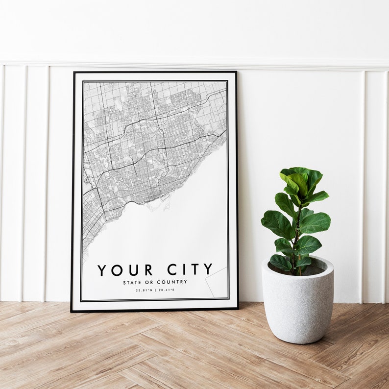May include: Black and white city map print in a black frame with the text "YOUR CITY STATE OR COUNTRY 22.81&deg;N | 90.41&deg;E" hanging on a white wall with a potted plant in front of it.
