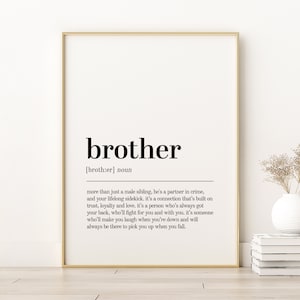 May include: A framed print featuring the word "brother" in a bold, black font, with a definition below. The print is in a gold frame and is displayed on a white wall. A vase with dried flowers and a stack of books are in the background.