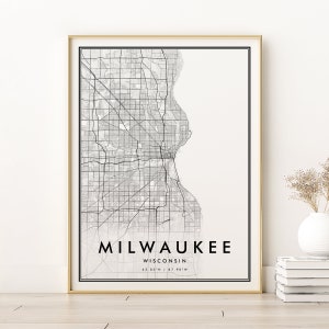 May include: A framed map print of Milwaukee, Wisconsin, in black and white. The city map is detailed with streets and the shoreline. The text "MILWAUKEE WISCONSIN" is centered at the bottom, with coordinates below. The frame is gold.