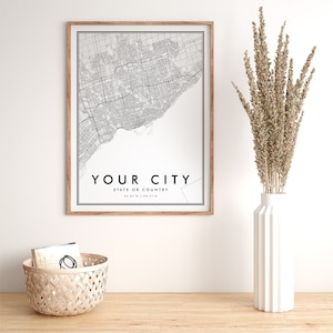 May include: A black and white city map print in a light wood frame. The map shows the streets of a city with the text "YOUR CITY" and "STATE OR COUNTRY" below the map. The print is hanging on a white wall above a wooden table.