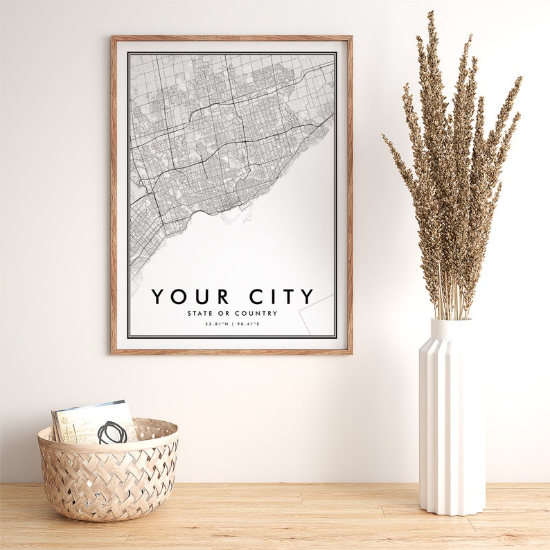 May include: A black and white city map print in a light wood frame. The map shows the streets of a city with the text "YOUR CITY" and "STATE OR COUNTRY" below the map. The print is hanging on a white wall above a wooden table.