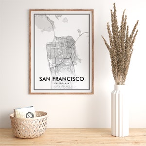 May include: A black and white map print of San Francisco, California in a wooden frame. The map shows the city streets and landmarks. The print is hanging on a white wall above a wooden table.