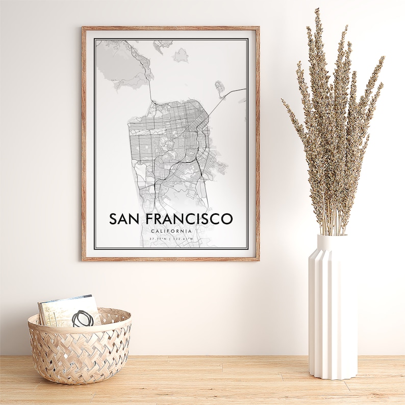 May include: A black and white map print of San Francisco, California in a wooden frame. The map shows the city streets and landmarks. The print is hanging on a white wall above a wooden table.