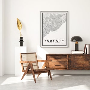 May include: A black and white city map print with the text "YOUR CITY" and "STATE OR COUNTRY" in a black frame hanging on a white wall above a wooden dresser with two drawers and two doors. A black table lamp with a mushroom-shaped shade is on the dresser. A wooden chair with a woven cane seat is in the foreground.