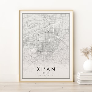 Op de afbeelding: Een zwart-witte kaartprint van Xi'an, China, met de naam van de stad en de coördinaten onder de kaart. De kaart is ingelijst in een gouden lijst en wordt getoond op een witte muur.