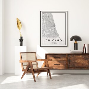May include: A black and white map print of Chicago, Illinois, with the city name and state printed below the map. The print is framed in a black frame and is hanging on a white wall. A wooden chair with a woven cane seat is in the foreground, and a wooden dresser with two drawers is in the background. A black lamp with a gold base is on the dresser.
