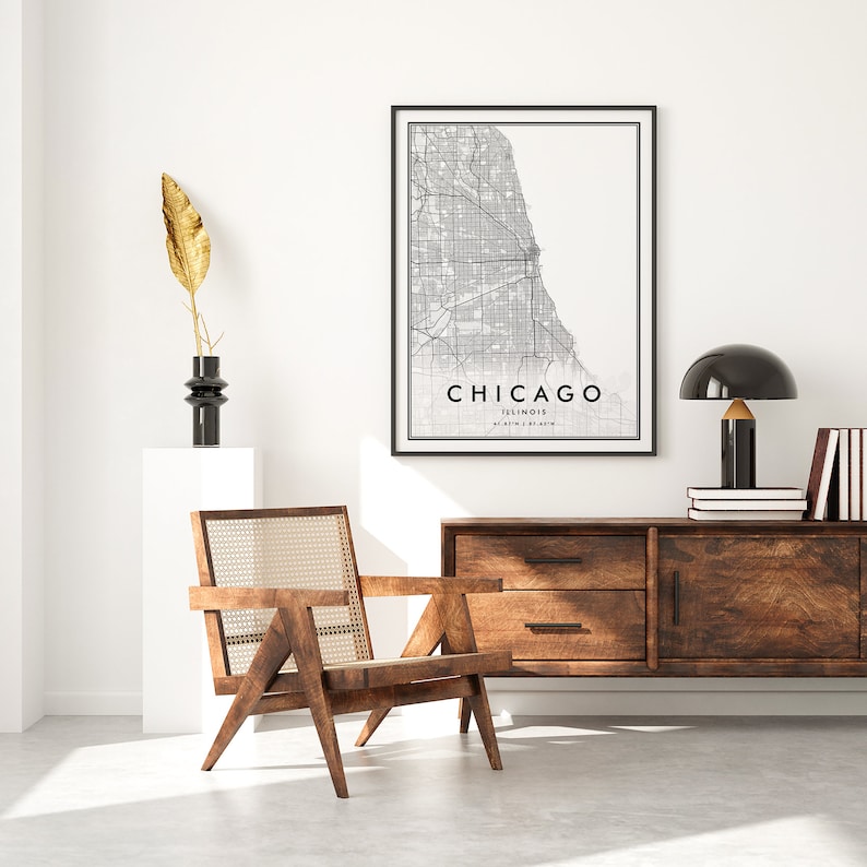 May include: A black and white map print of Chicago, Illinois, with the city name and state printed below the map. The print is framed in a black frame and is hanging on a white wall. A wooden chair with a woven cane seat is in the foreground, and a wooden dresser with two drawers is in the background. A black lamp with a gold base is on the dresser.
