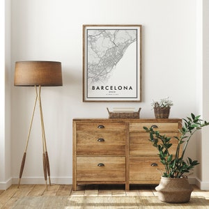 May include: A framed black and white map print of Barcelona, Spain, hanging on a white wall above a wooden dresser with six drawers. The dresser has black metal drawer pulls. A brown lamp with a tripod stand is on the left side of the image. A potted plant is on the right side of the image.