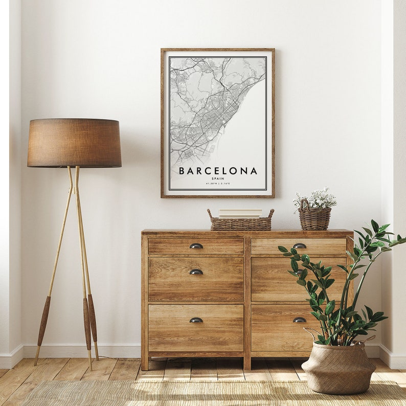 May include: A framed black and white map print of Barcelona, Spain, hanging on a white wall above a wooden dresser with six drawers. The dresser has black metal drawer pulls. A brown lamp with a tripod stand is on the left side of the image. A potted plant is on the right side of the image.