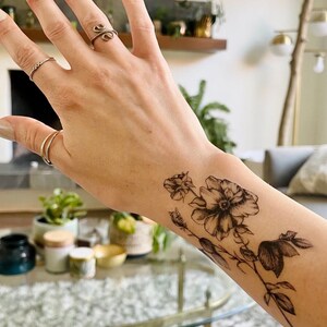 May include: A black and white temporary tattoo of a floral design with three flowers and leaves on a person's wrist.