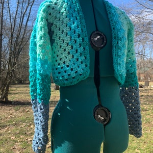 Puede incluir: Un cárdigan corto de ganchillo con mangas largas en tonos azules y verdes.