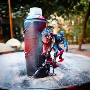 May include: A spray paint can with a red, blue, and black paint splatter design. A small action figure of Captain America is standing on top of the can.