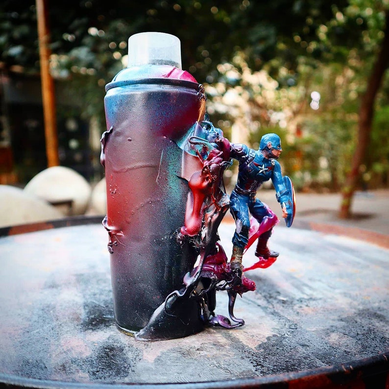 May include: A spray paint can with a red, blue, and black paint splatter design. A small action figure of Captain America is standing on top of the can.