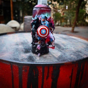 May include: A spray paint can with a red, white, and blue superhero figure attached to it. The figure is holding a shield with a star in the center. The can is sitting on a red and black surface.