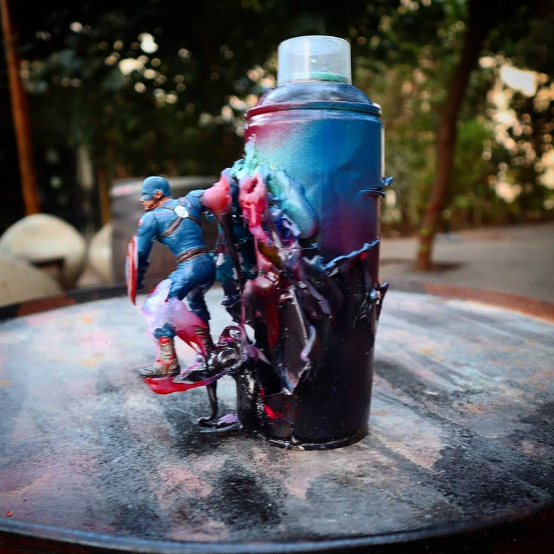May include: A blue and red spray paint can with a superhero action figure standing on top of it. The can is covered in drips and splatters of paint.