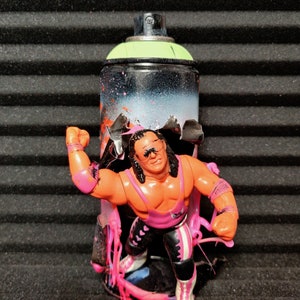 May include: A pink and black spray paint can with a wrestler action figure inside. The can is covered in pink paint splatters and the wrestler is wearing a pink and black wrestling outfit.
