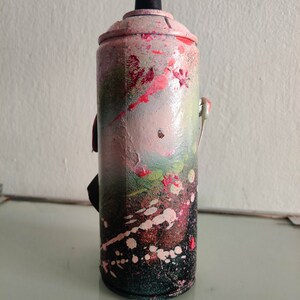 Puede incluir: Una lata de pintura en aerosol rosa y verde con detalles negros y salpicaduras de pintura blanca. La lata tiene una tapa negra y una leng&uuml;eta de apertura plateada.