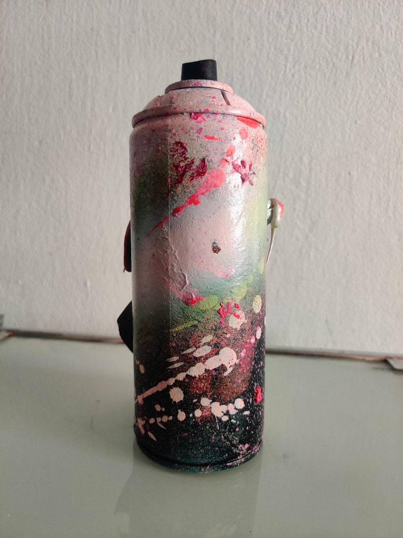 Puede incluir: Una lata de pintura en aerosol rosa y verde con detalles negros y salpicaduras de pintura blanca. La lata tiene una tapa negra y una leng&uuml;eta de apertura plateada.