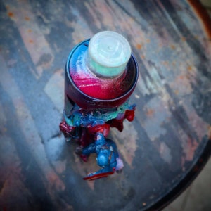 May include: A red, blue, and green spray paint can with a white cap. The can is partially covered in a blue and red plastic action figure.
