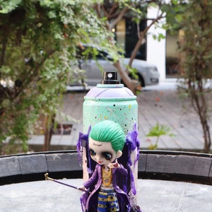 Puede incluir: Una figura decorativa de un personaje con cabello verde, abrigo morado y cadena dorada, sosteniendo un bastón, frente a una lata de pintura en aerosol turquesa y morada. La palabra "Damaged" es visible en la frente del personaje.