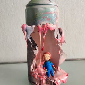 Puede incluir: Una lata de pintura en aerosol con la parte superior y los lados rotos, revelando una sustancia pegajosa rosa y roja en el interior. Una peque&ntilde;a figura de juguete de pl&aacute;stico de un ni&ntilde;o con cabello rojo est&aacute; de pie dentro de la lata.