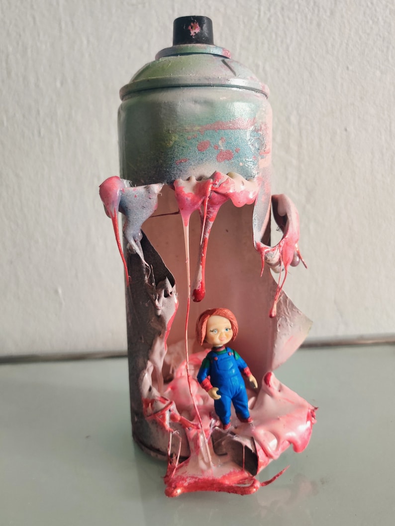 Puede incluir: Una lata de pintura en aerosol con la parte superior y los lados rotos, revelando una sustancia pegajosa rosa y roja en el interior. Una peque&ntilde;a figura de juguete de pl&aacute;stico de un ni&ntilde;o con cabello rojo est&aacute; de pie dentro de la lata.