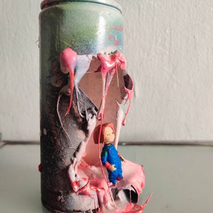 Puede incluir: Una lata de pintura en aerosol con un gran agujero en el lateral, que revela una sustancia pegajosa rosa y roja. Una peque&ntilde;a figura de mu&ntilde;eca de pl&aacute;stico est&aacute; de pie dentro de la lata, mirando hacia afuera del agujero.