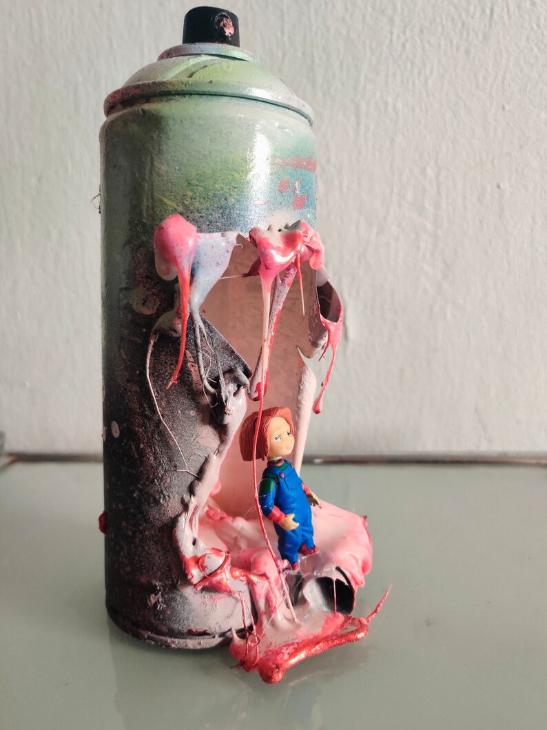 Puede incluir: Una lata de pintura en aerosol con un gran agujero en el lateral, que revela una sustancia pegajosa rosa y roja. Una peque&ntilde;a figura de mu&ntilde;eca de pl&aacute;stico est&aacute; de pie dentro de la lata, mirando hacia afuera del agujero.