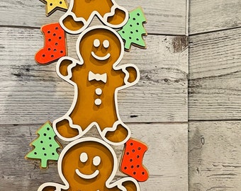 May include: A stack of three gingerbread men ornaments with colorful accents, including stars, trees, and stockings. The ornaments are stacked on a wooden base with a white bead garland.
