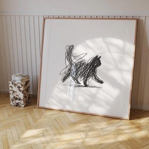 May include: A framed art print depicting a black scribble drawing of a cat. The artwork is set against a white background, framed in light wood. The print is displayed on a wall above a wooden floor.