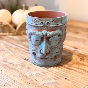 Puede incluir: Una maceta de cerámica azul y marrón con un diseño de cara de mono. La maceta tiene un acabado envejecido, con pintura azul sobre una base marrón. La cara del mono presenta ojos cerrados y una boca sonriente. El interior es de color marrón liso.