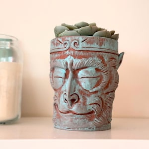 May include: A terracotta planter shaped like a monkey's face with a green glaze. The planter has a small succulent plant growing inside.