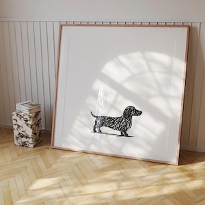以下が含まれることがあります： ダックスフンドの白黒イラストが描かれた、額入りのアートプリント。犬は白い背景に落書き風に描かれています。フレームは薄茶色で、木製の床に飾られています。