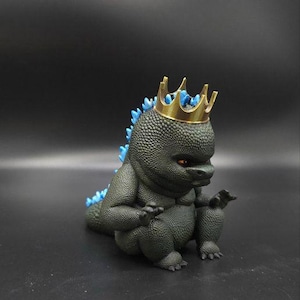May include: A dark gray figurine of a Godzilla-like creature wearing a gold crown. The figurine has blue spikes down its back and orange eyes. The creature is in a seated position with its hands resting in front.